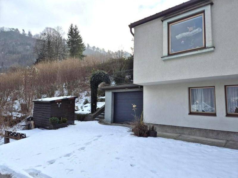 Seitenansicht des Hauses mit Garage, kleinem Holzschuppen und verschneitem Hang im Hintergrund.