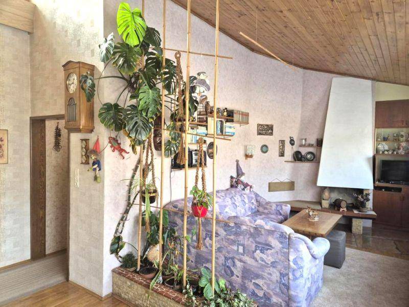 Geräumiges Wohnzimmer mit Holzbalkendecke, Kamin und vielen Pflanzen als Raumteiler.