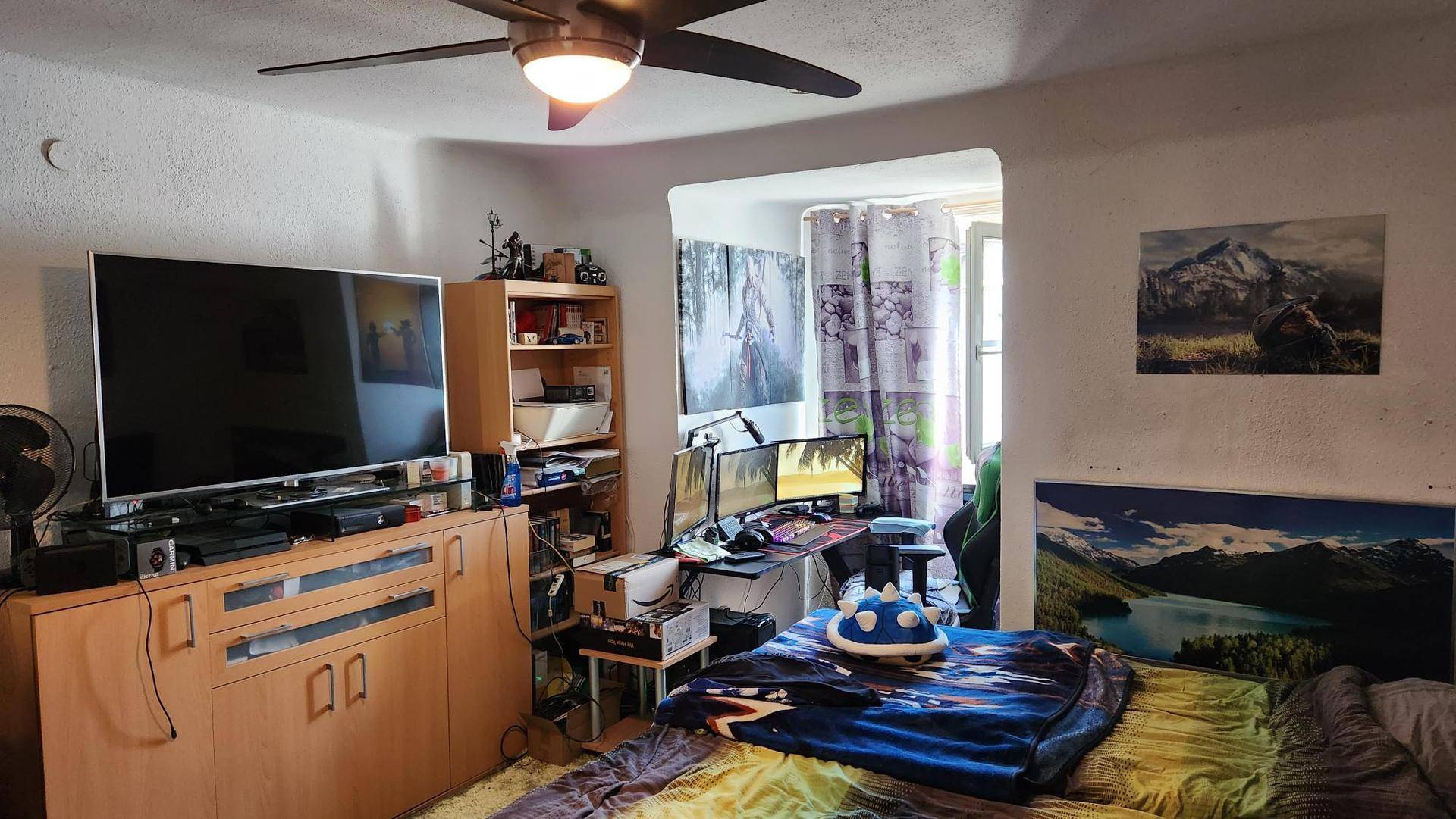 Schlafzimmer mit Gaming-Setup, großem Fernseher und Deckenventilator.