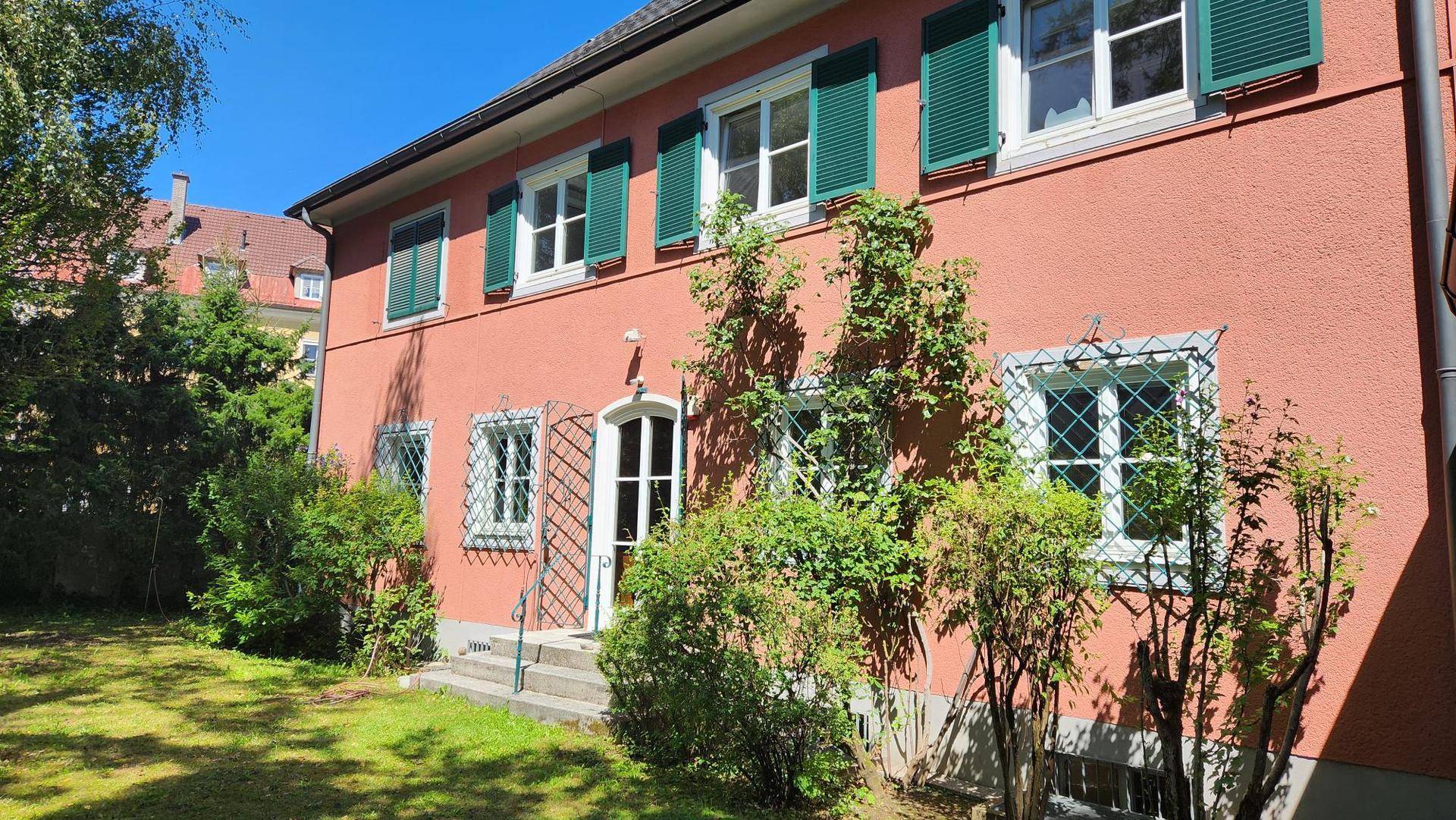 Ansicht des roten Gebäudes vom Garten aus mit grünen Fensterläden und üppiger Vegetation.