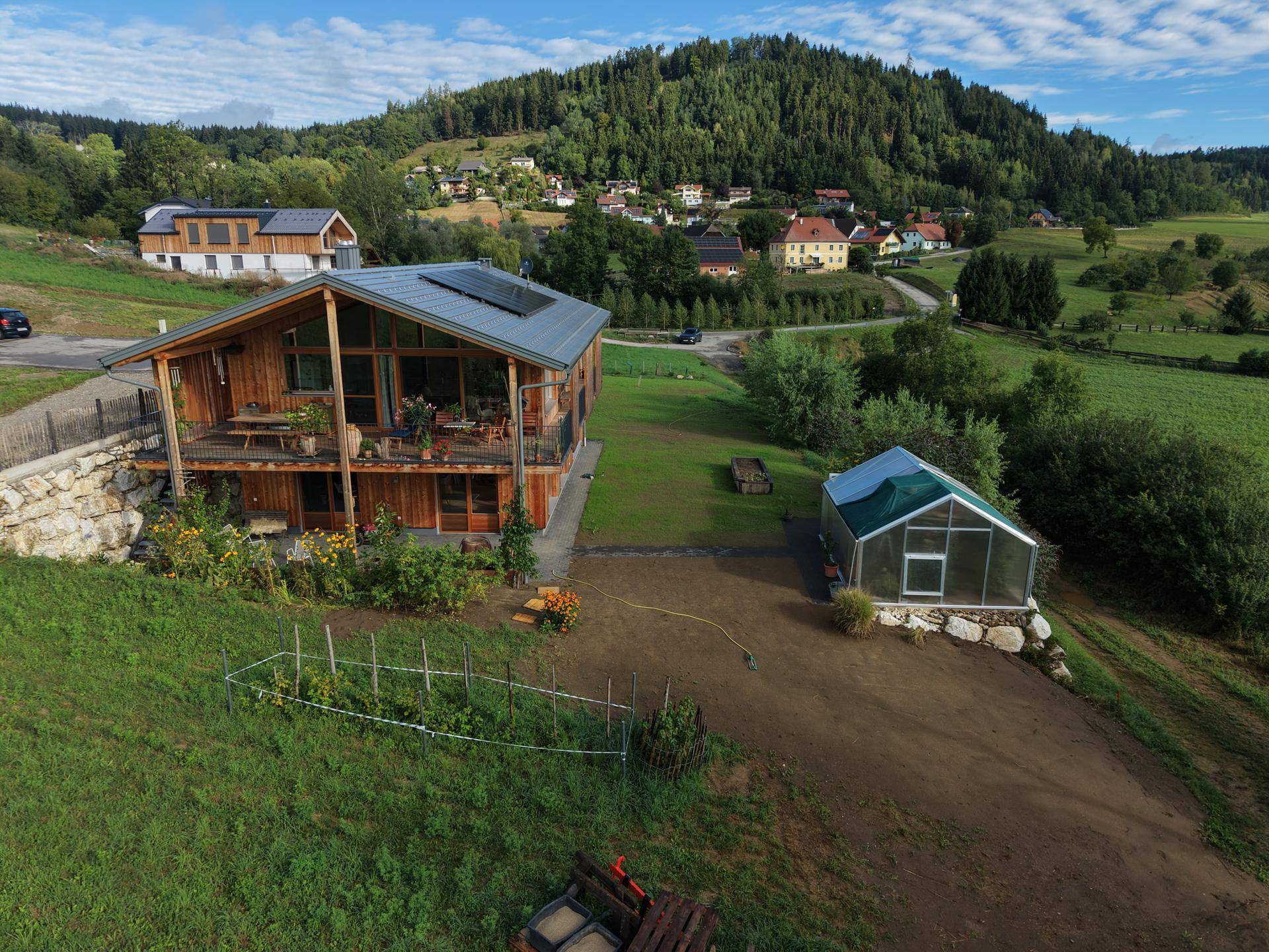 Holzhaus mit Balkon und Gewächshaus in ländlicher Umgebung, umgeben von grünen Hügeln.