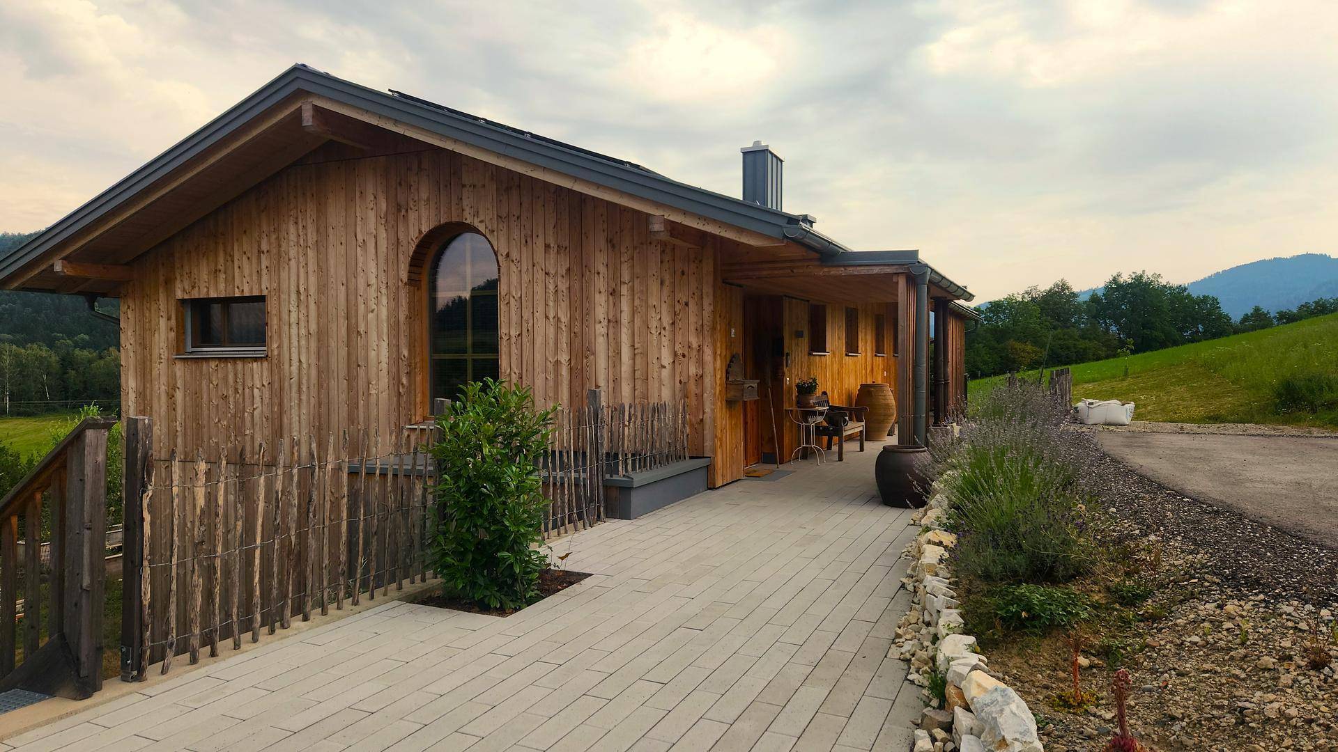 Holzhaus mit gepflastertem Weg und Blick auf die umliegende grüne Landschaft unter bewölktem Himmel.