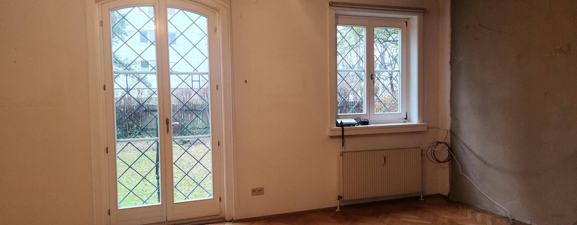 Heller Raum mit Fischgrätparkett, einer großen Fenstertür und einem weiteren Fenster mit Heizkörper.