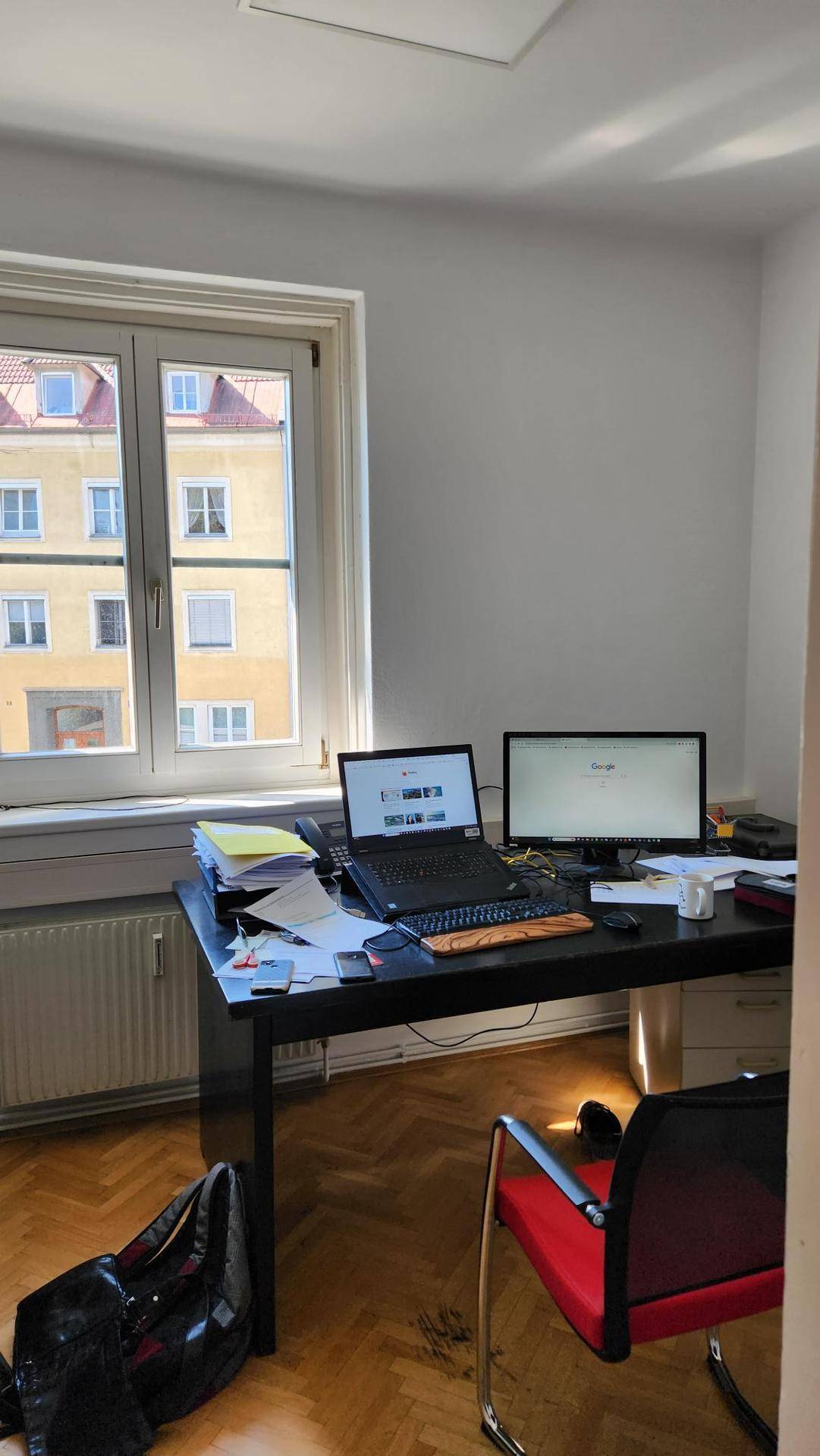 Helles Büro mit Schreibtisch, Computern und Blick auf die umliegenden Gebäude durch das Fenster.