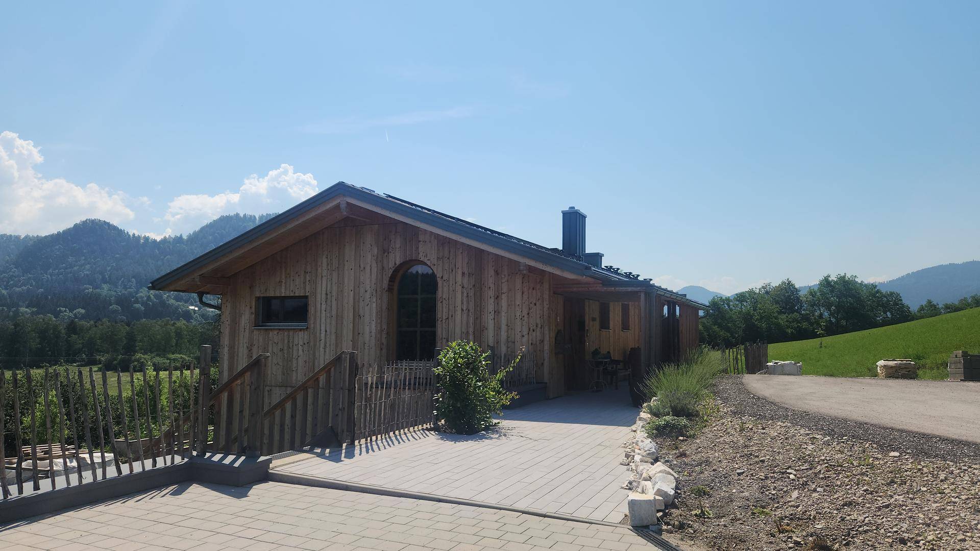 Holzhaus mit gepflastertem Vorplatz und Blick auf die umliegende grüne Hügellandschaft.