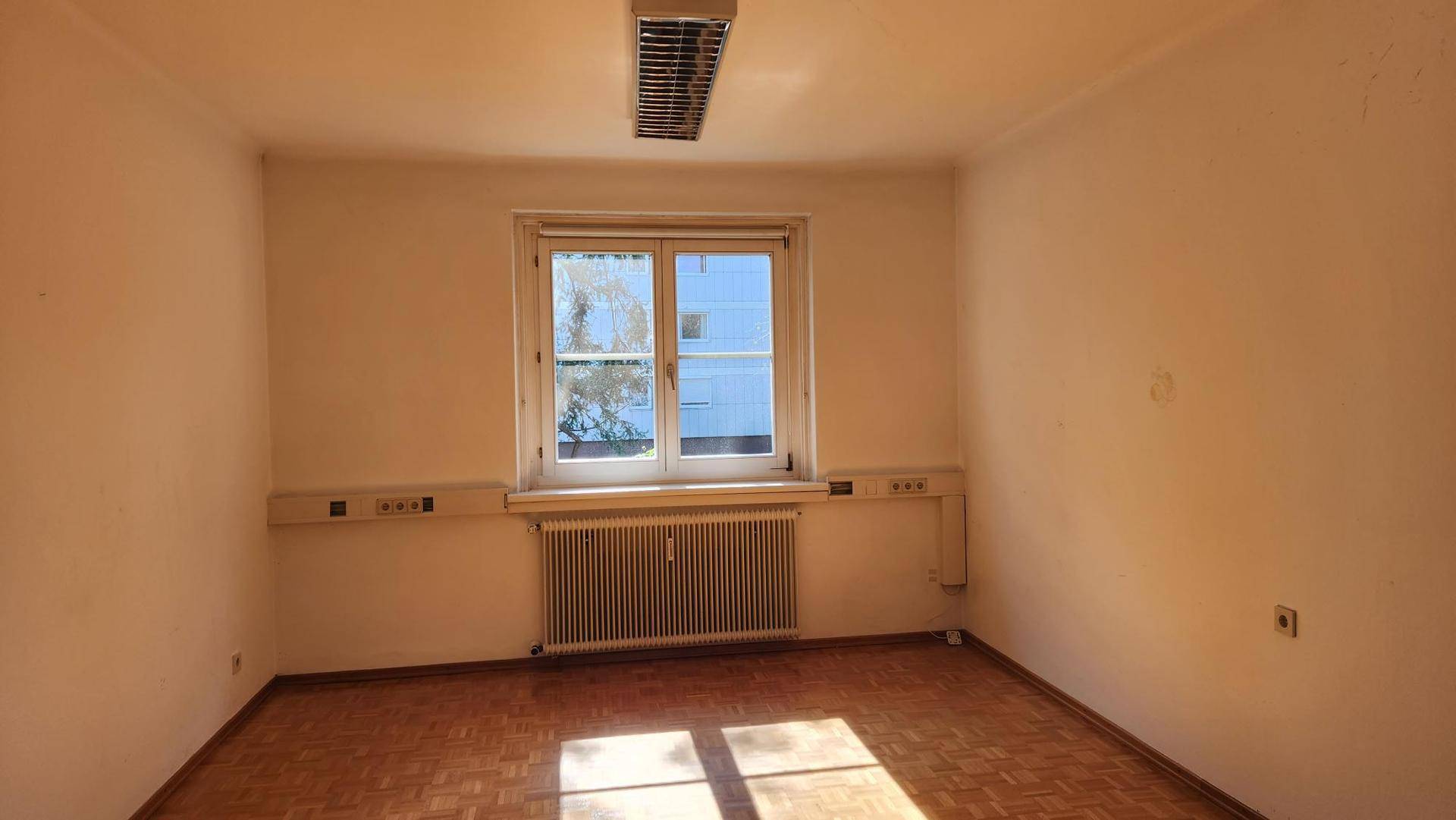 Leerer Büroraum mit Holzboden, einem Fenster und einer Deckenleuchte, bereit zur Einrichtung.