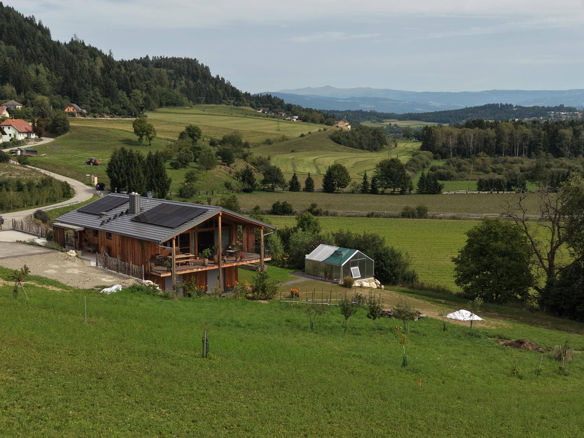 Holzhaus mit Solaranlage auf dem Dach, umgeben von weiten grünen Feldern und Hügeln.