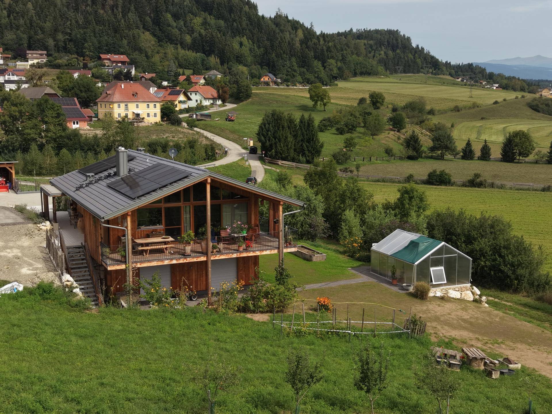 Modernes Holzhaus mit Gewächshaus und Solaranlage, eingebettet in eine ländliche Hügellandschaft.