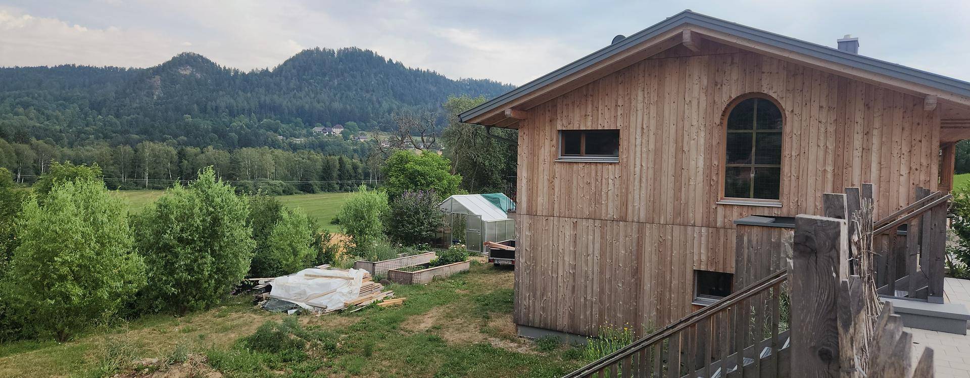 Holzhaus mit Hochbeeten und Gartenbereich, umgeben von grüner Natur und Hügeln.