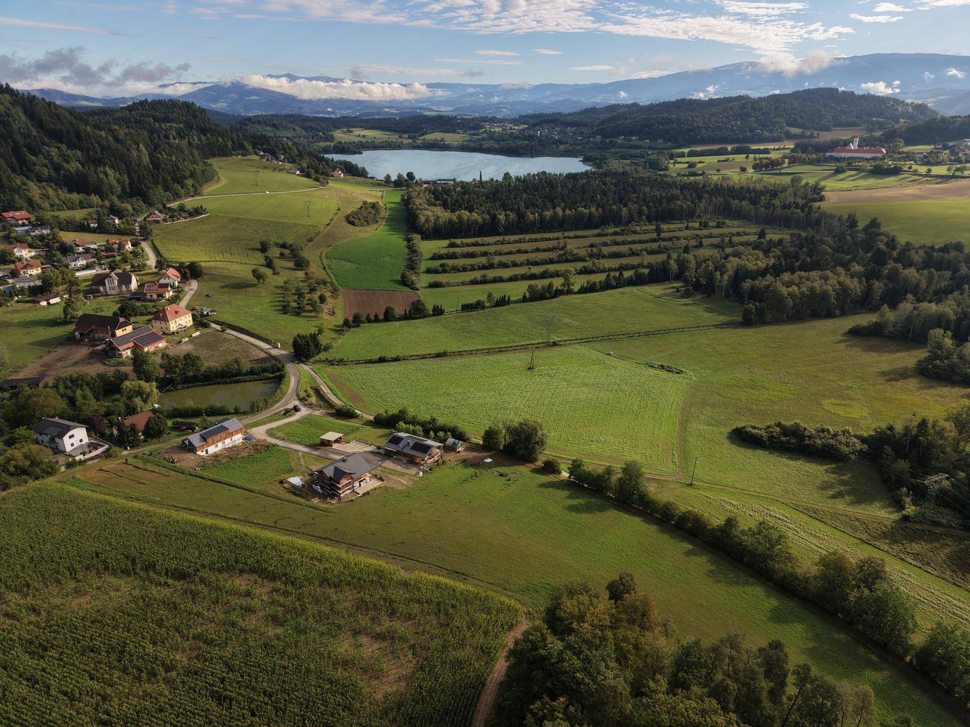 Weitläufige Landschaft mit See, grünen Feldern und vereinzelten Häusern unter blauem Himmel.