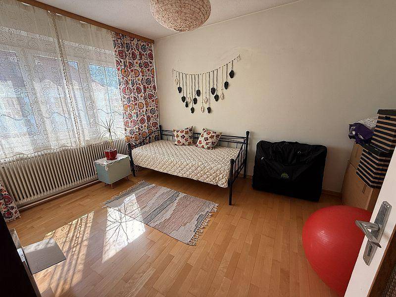 Helles Schlafzimmer mit Holzboden, Einzelbett und großem Fenster mit Vorhängen.