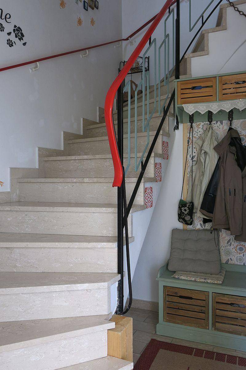 Heller Treppenaufgang mit beigefarbenen Stufen und einem auffälligen roten Handlauf, ergänzt durch eine Garderobe.