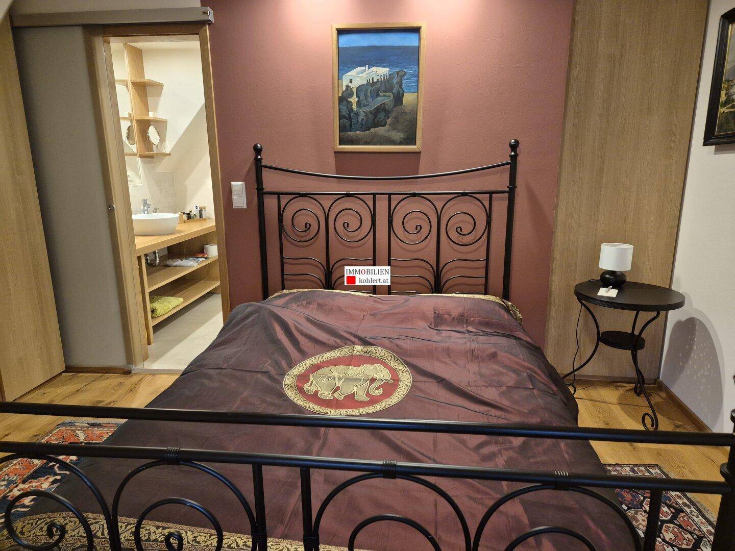 Schlafzimmer mit schmiedeeisernem Bett, roter Wand und Zugang zu einem angrenzenden Bad.