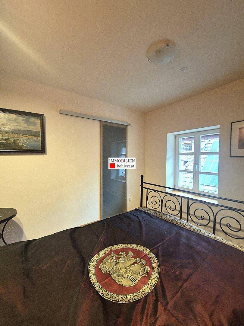 Schlafzimmer mit Holzboden, Fenster und Schiebetür, die zu einem weiteren Raum führt.