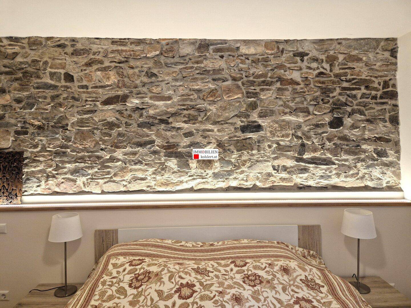 Schlafzimmer mit einer markanten Steinwand als Akzent und indirekter Beleuchtung über dem Bett.