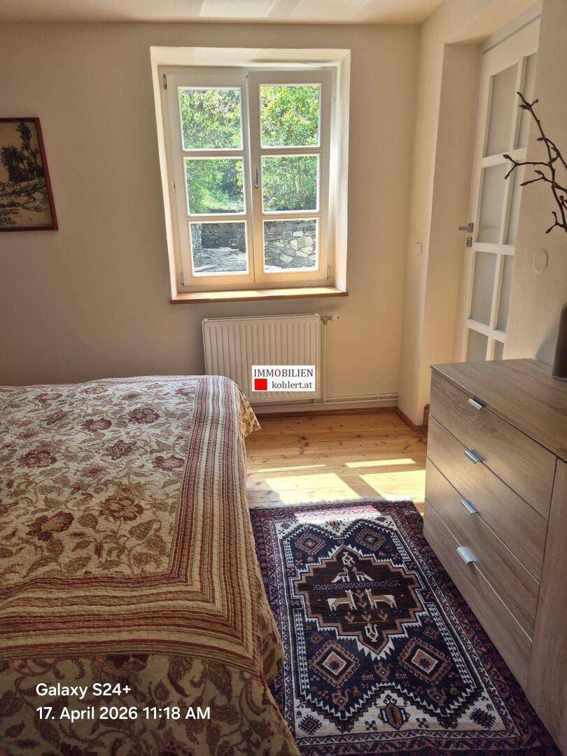 Helles Schlafzimmer mit Holzdielenboden, Fenster und einem Bett mit gemusterter Decke.