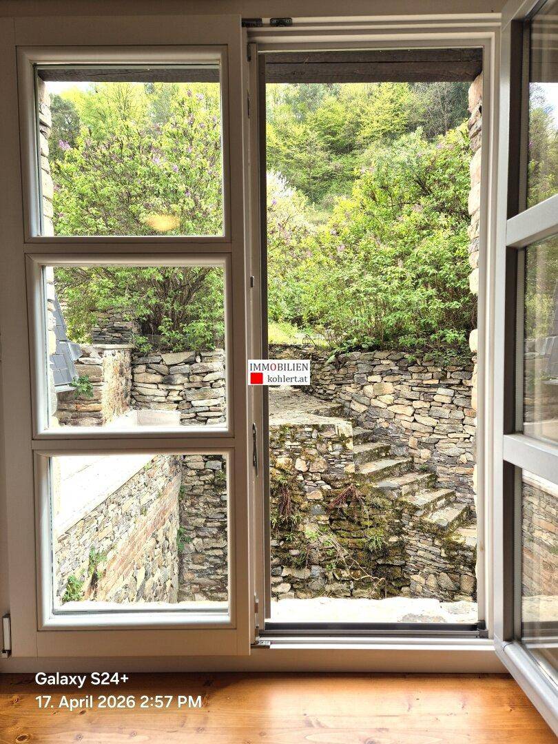 Blick aus dem Fenster auf einen terrassierten Garten mit Steinmauern und üppiger Vegetation.