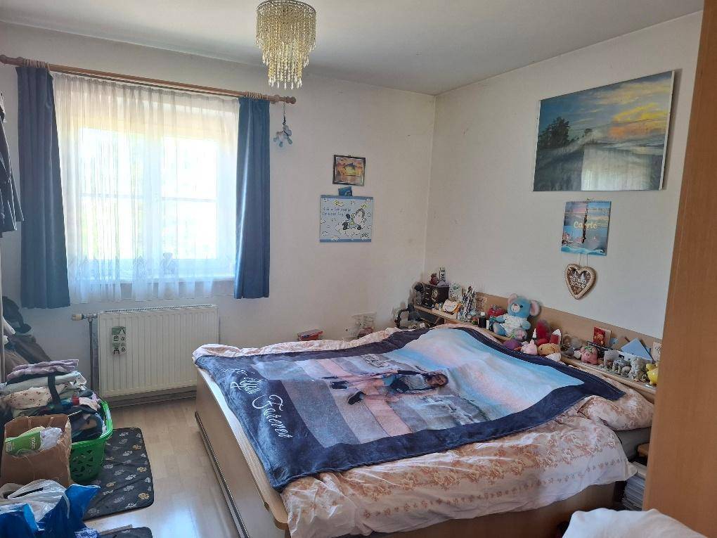 Schlafzimmer mit einem Bett, einem großen Fenster, Heizkörper und einem Kronleuchter an der Decke.