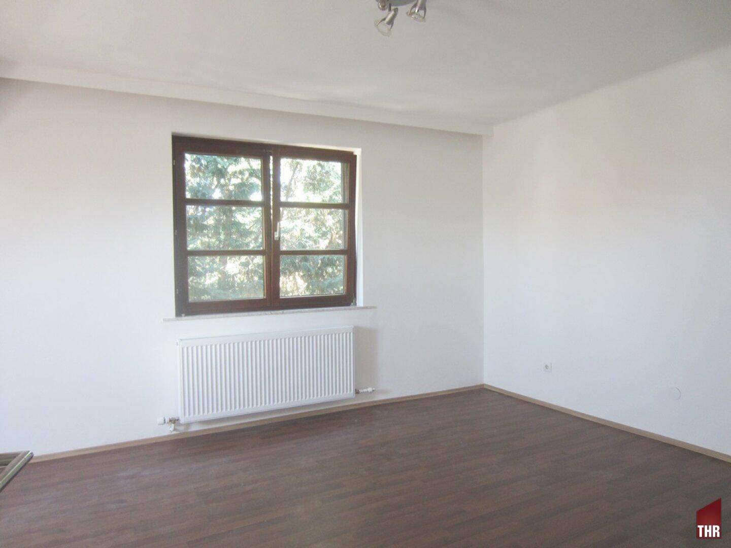 Einfaches, helles Zimmer mit Laminatboden, großem Fenster und Heizkörper unter dem Fenster.