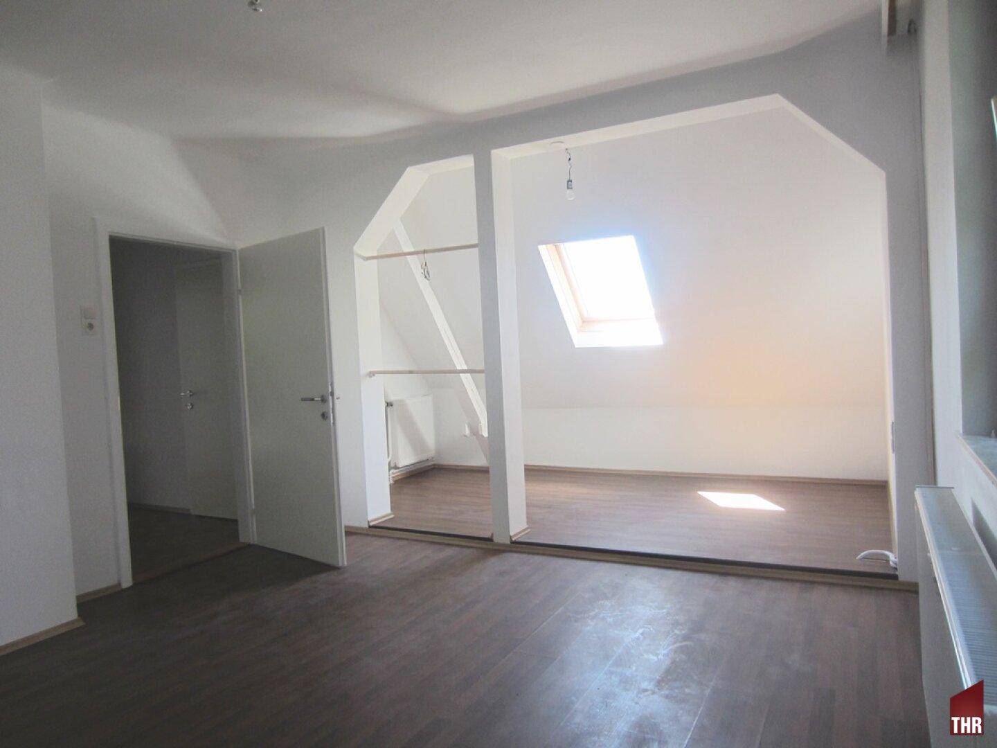 Helles Dachgeschosszimmer mit Laminatboden, Dachfenster und offenem Übergang zu einem Nebenbereich.