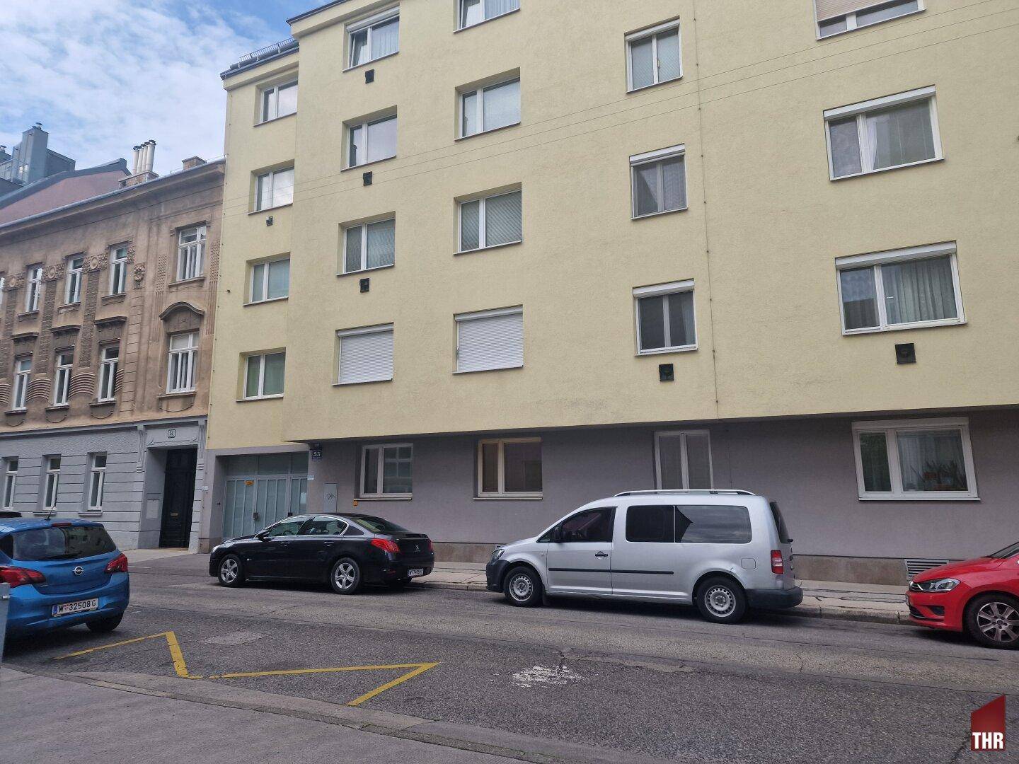 Mehrfamilienhaus mit heller Fassade und parkenden Autos auf der Straße, typisch für eine städtische Umgebung.