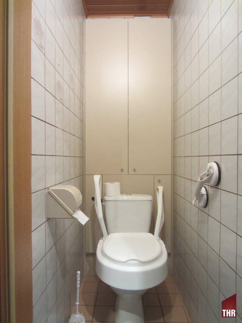 Separate Toilette mit weißen Fliesen, WC und praktischen Ablageschränken, ausgestattet mit Haltegriffen.