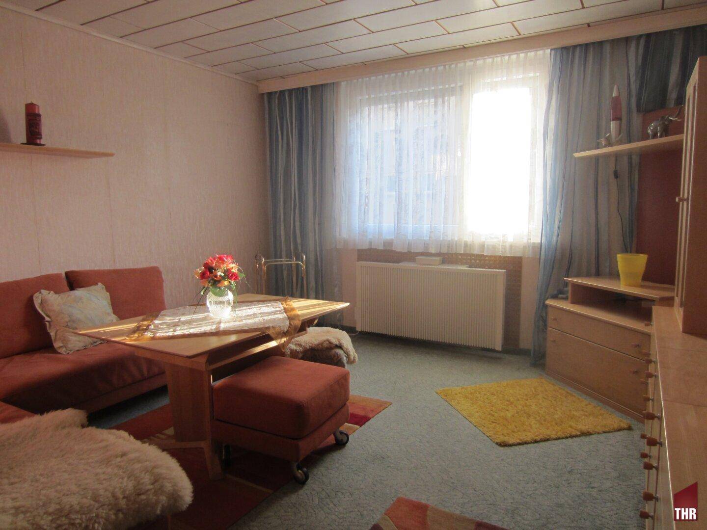 Helles Wohnzimmer mit Teppichboden, gemütlicher Sitzecke und großem Fenster, das viel Tageslicht hereinlässt.