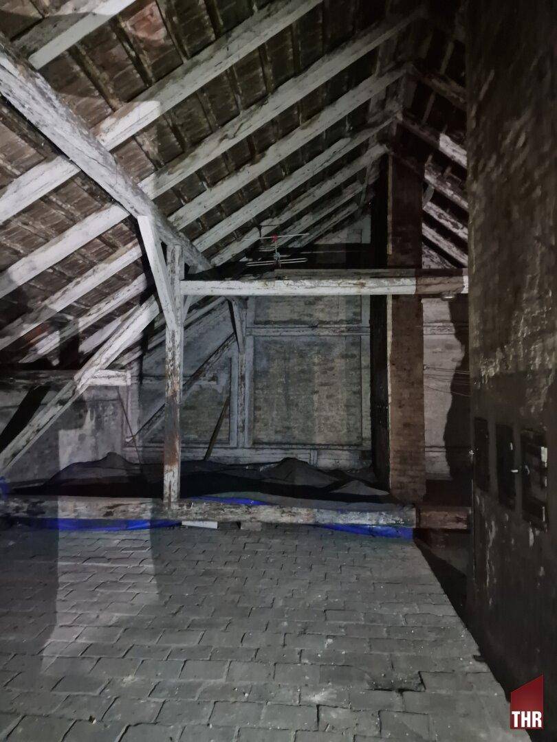 Dunkler Dachboden mit sichtbaren Holzbalken und Ziegelboden, der das alte Dachgebälk zeigt.