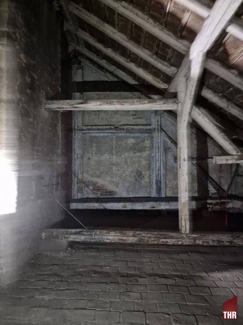 Dachboden mit sichtbaren Holzbalken, Ziegelboden und einem kleinen Fenster, das etwas Licht hereinlässt.