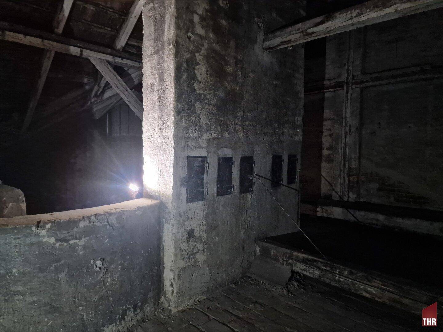 Weiterer Blick in den dunklen Dachboden mit einer Ziegelwand und mehreren alten Metallklappen.
