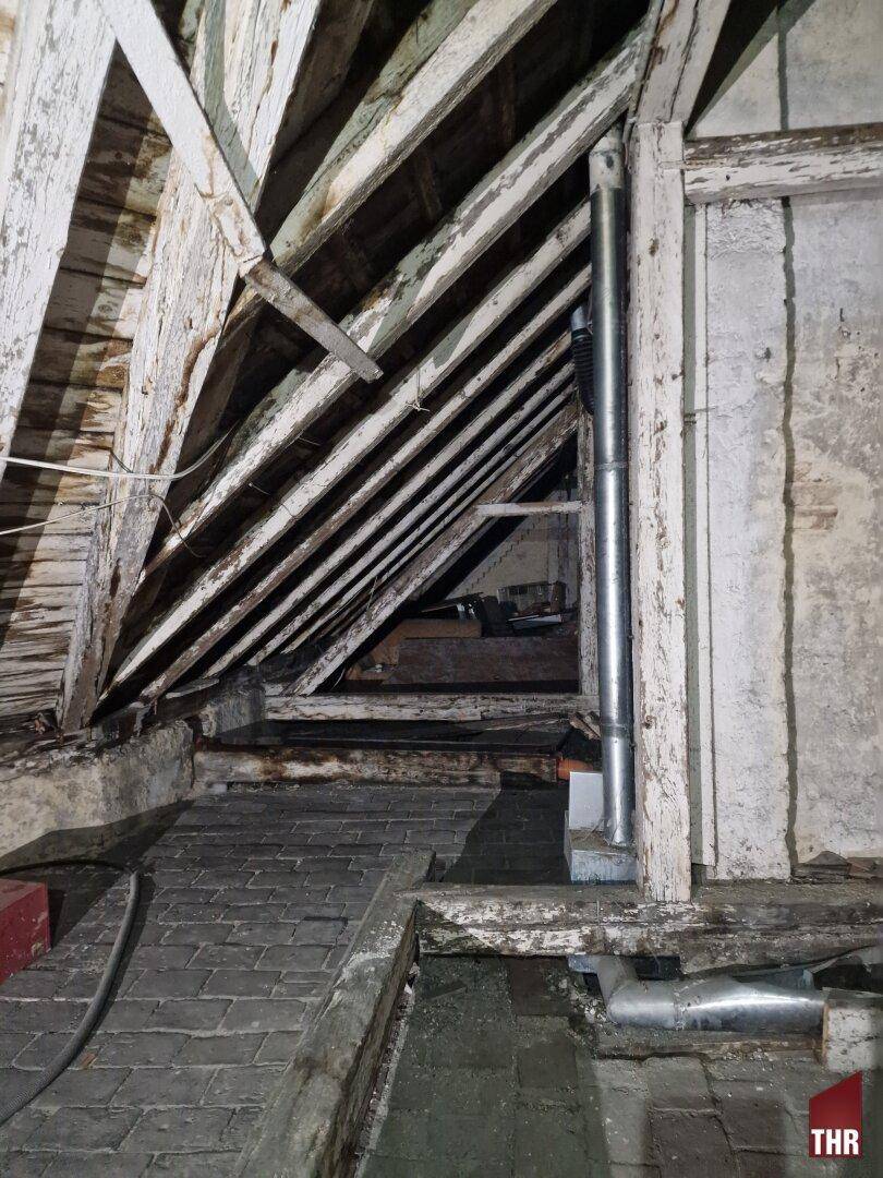 Dachboden mit freiliegenden Holzbalken, Ziegelboden und einer vertikal verlaufenden Metallleitung.