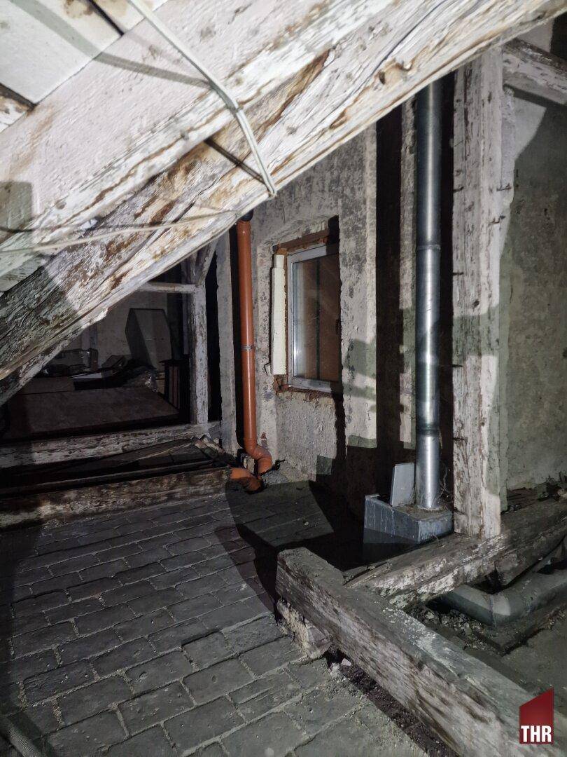 Dachboden mit Holzbalken, Ziegelboden, einem kleinen Fenster und sichtbaren Rohrleitungen.