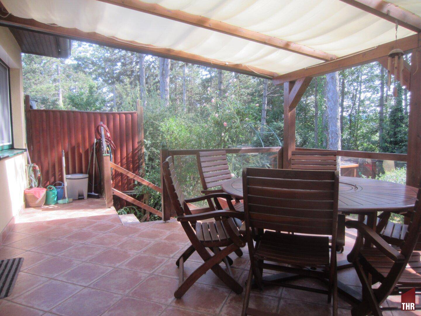 Überdachte Terrasse mit Holztisch und Stühlen, umgeben von Bäumen und einem Holzzaun.