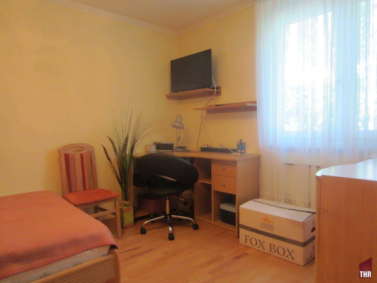 Helles Schlafzimmer mit Holzboden, Schreibtisch und großem Fenster mit viel Tageslicht.