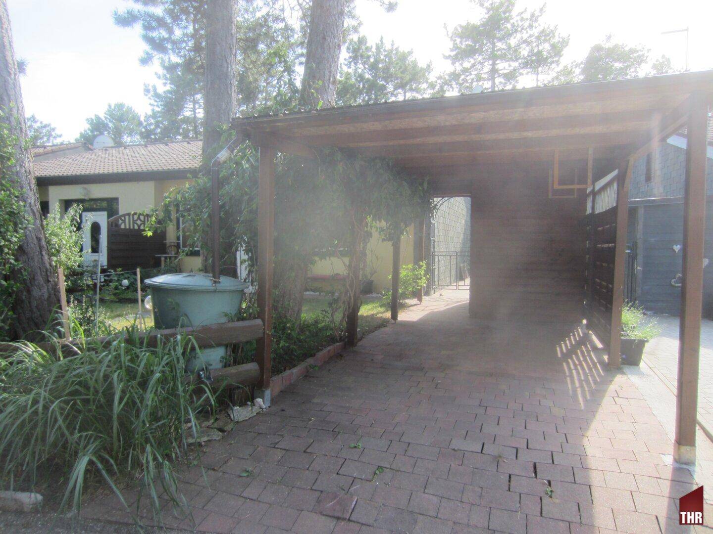Überdachter Stellplatz oder Carport mit gepflastertem Boden und umgebender Vegetation.