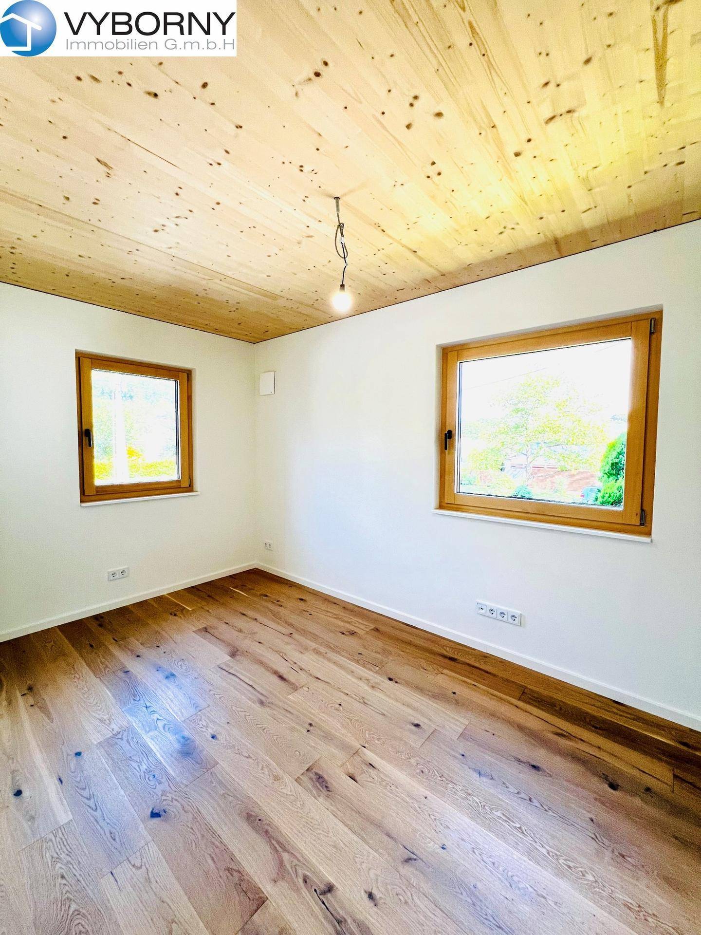 Helles Schlafzimmer mit Holzboden, Holzdecke und zwei Fenstern mit Blick ins Grüne.