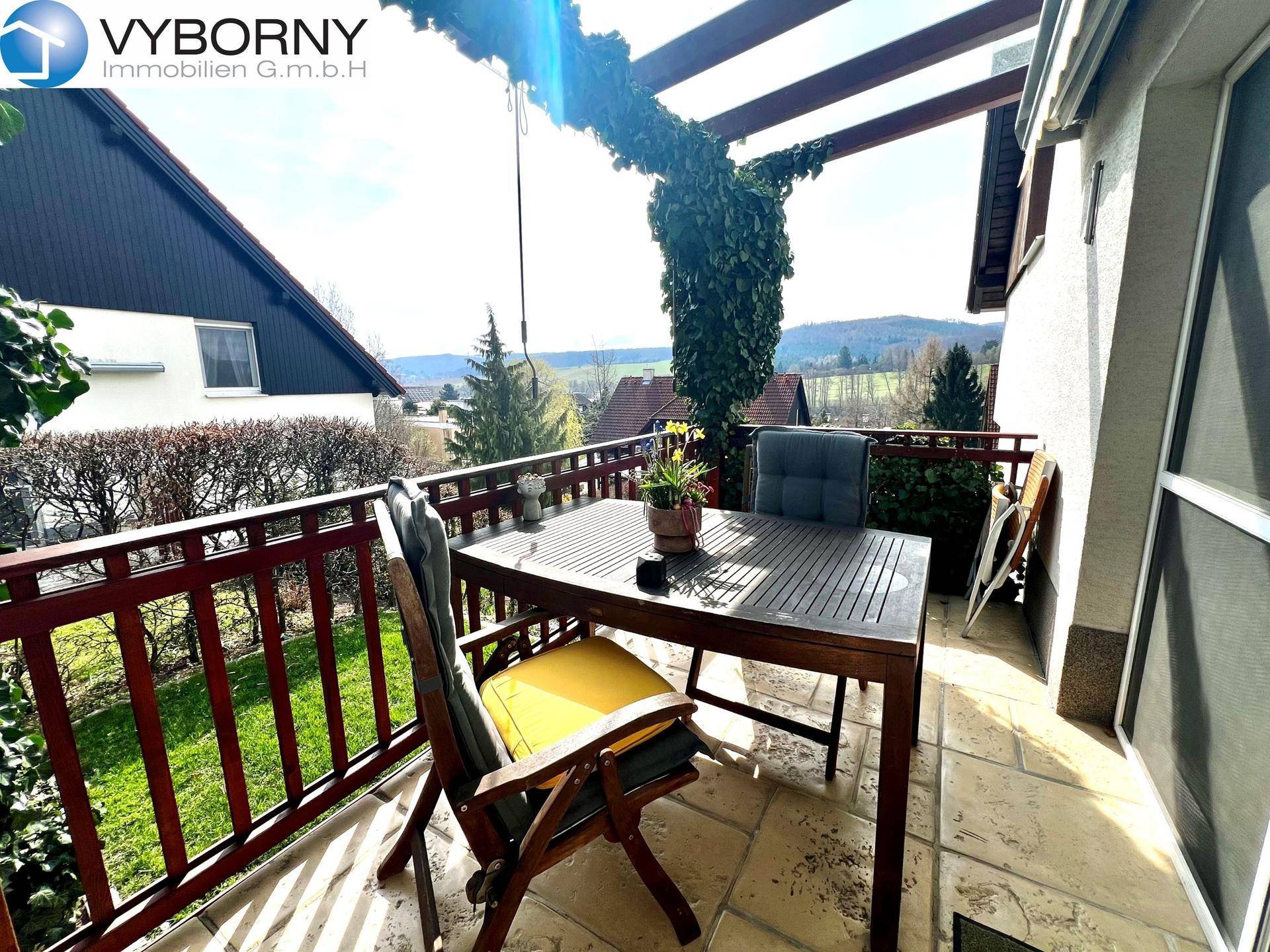 Möblierter Balkon mit Holztisch und Stühlen, umgeben von Grün und mit Fernblick.