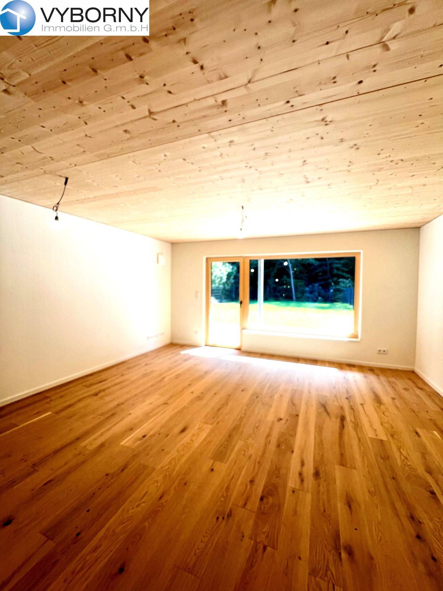 Heller Raum mit Holzboden, Holzdecke und großer Fensterfront mit Zugang zum Garten.