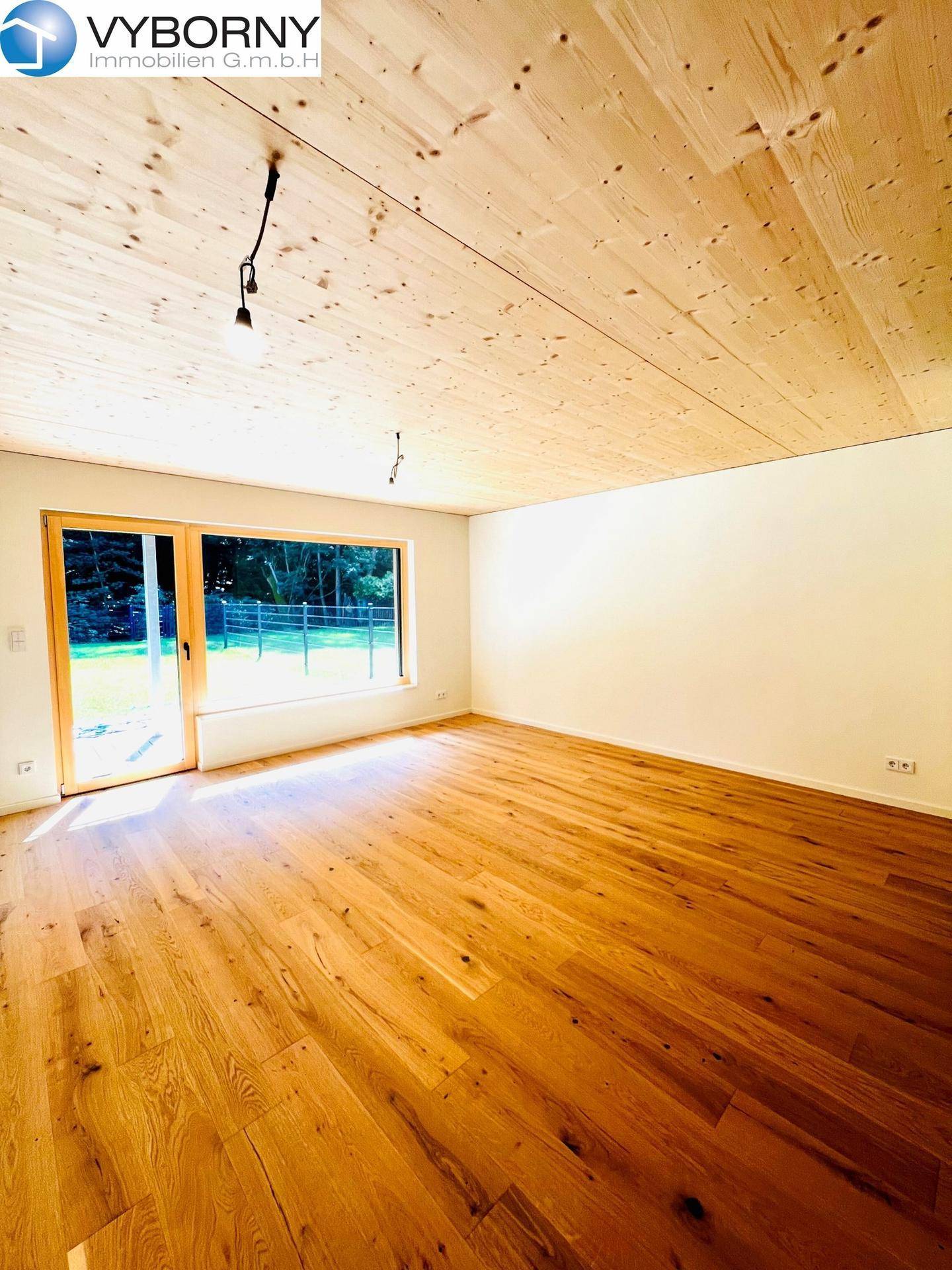 Lichtdurchfluteter Wohnraum mit Holzboden, Holzdecke und breiter Fensterfront zum Garten.