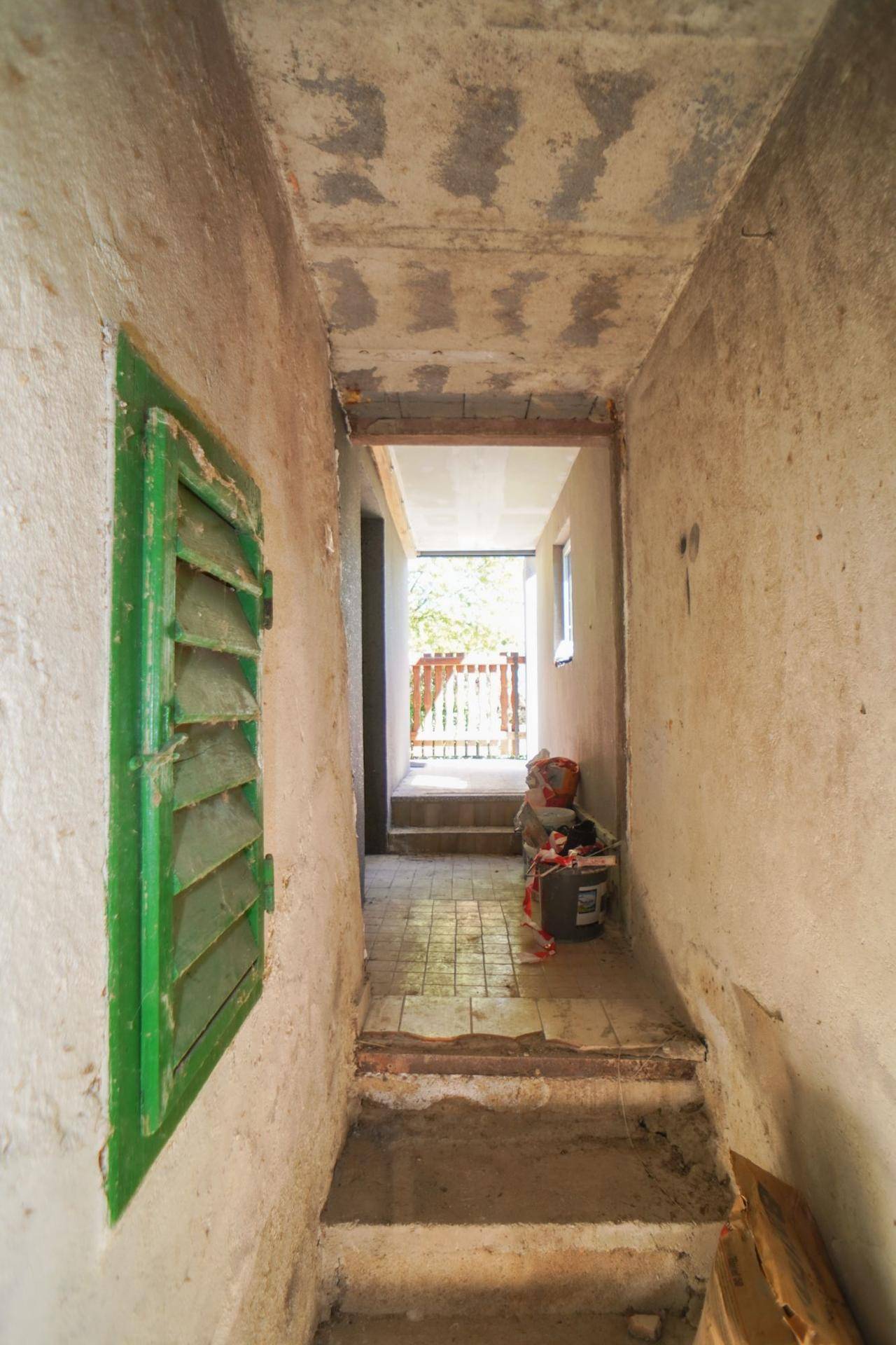 Alter Flur mit Treppen und einem Fenster mit grünen Fensterläden, renovierungsbedürftig.