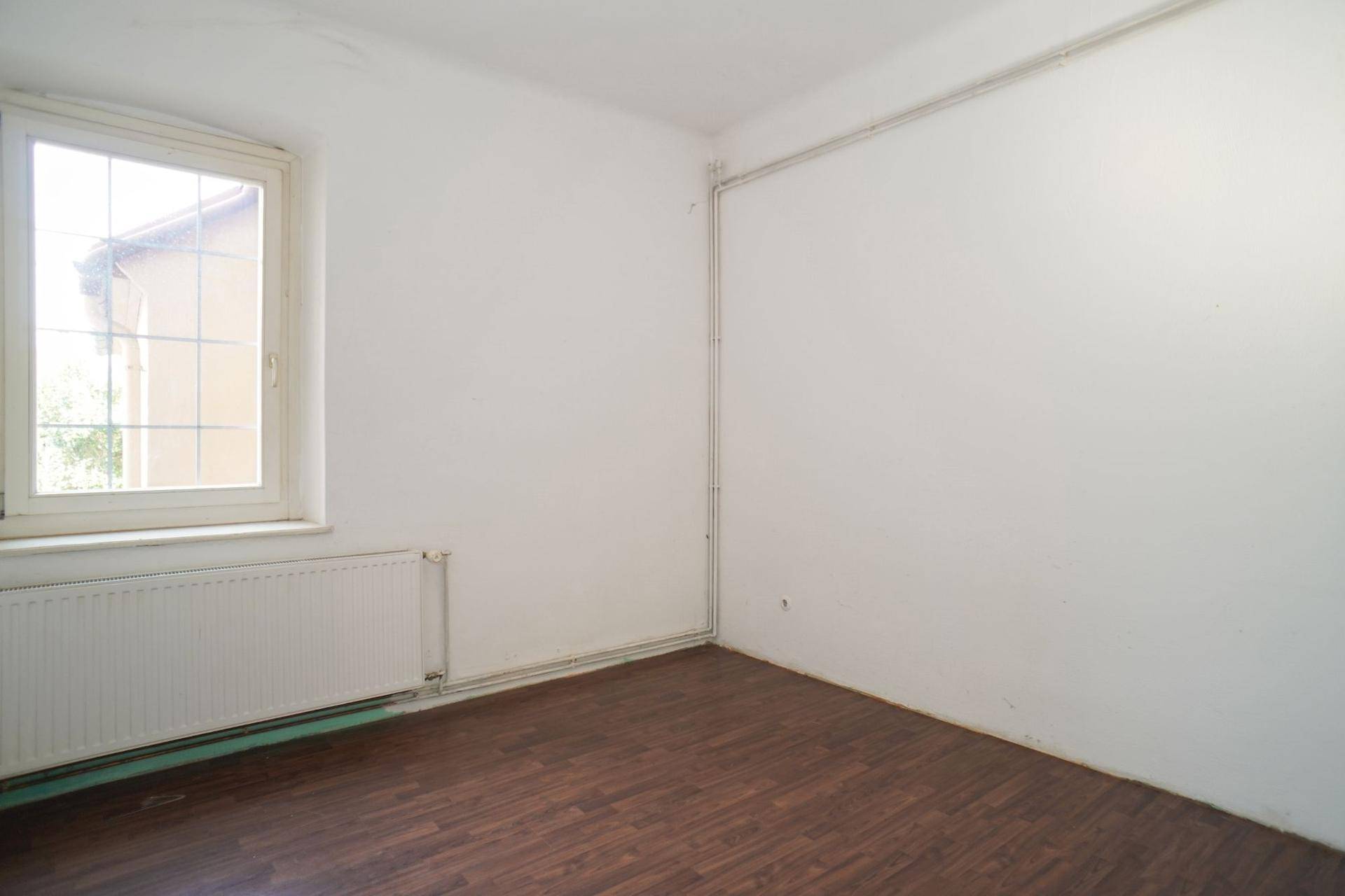 Einfacher, heller Raum mit weiß gestrichenen Wänden und dunklem Laminatboden, mit Fenster.
