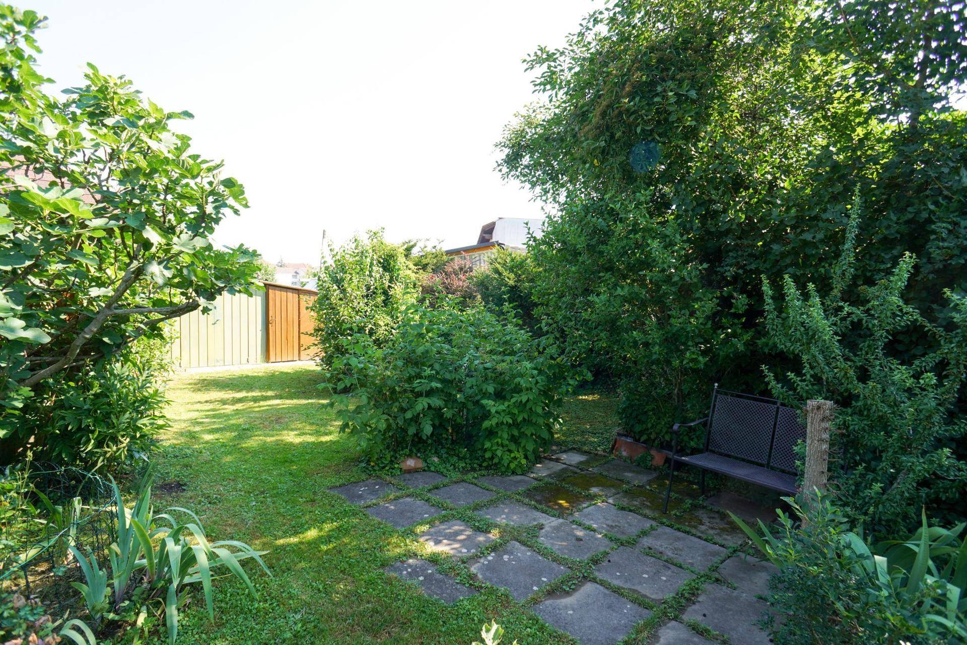 Großer Gartenbereich mit Rasenfläche, Steinplatten und einem Gartenhaus im Hintergrund.