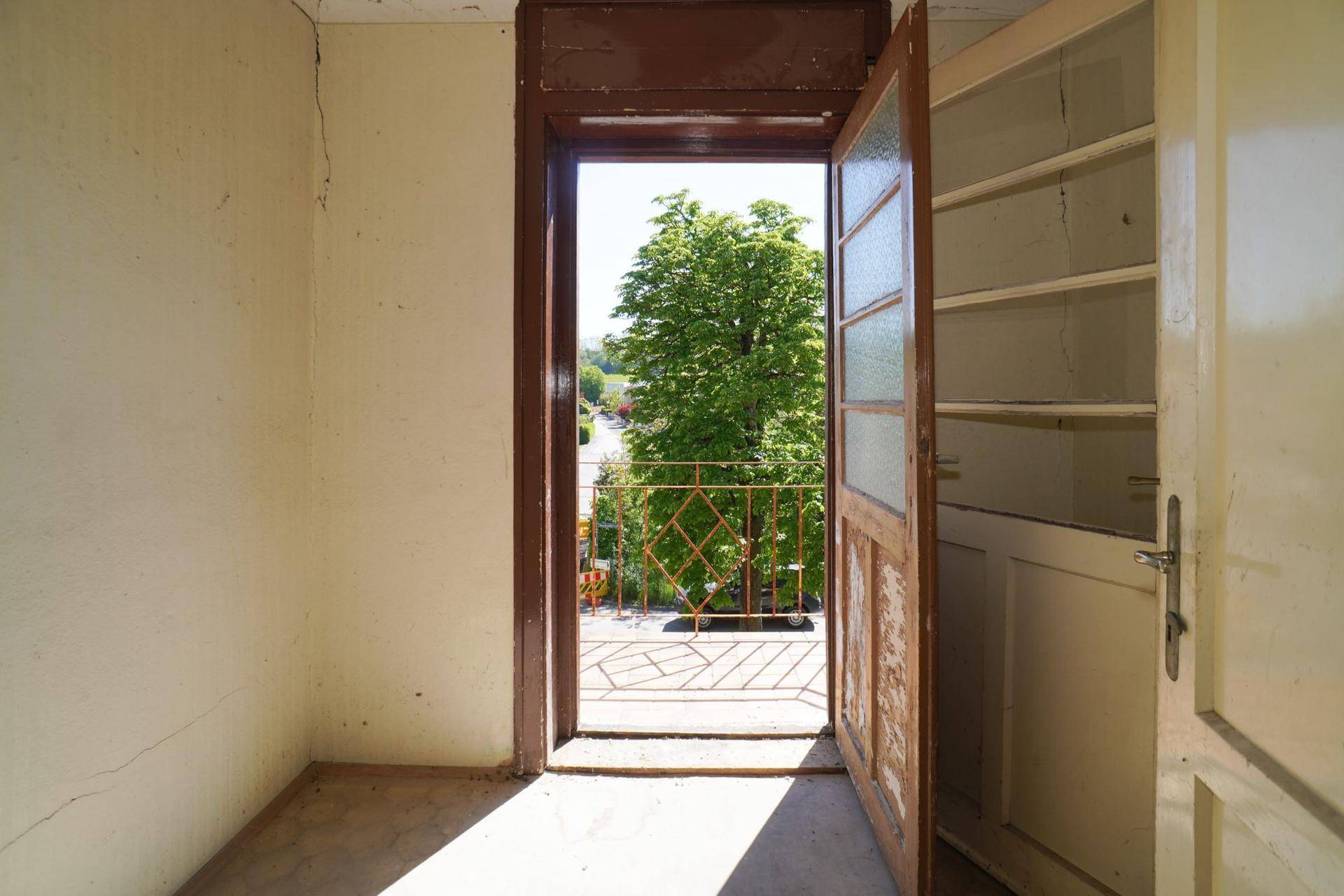 Blick von einem Innenraum durch eine offene Tür auf einen kleinen Balkon mit Baum.