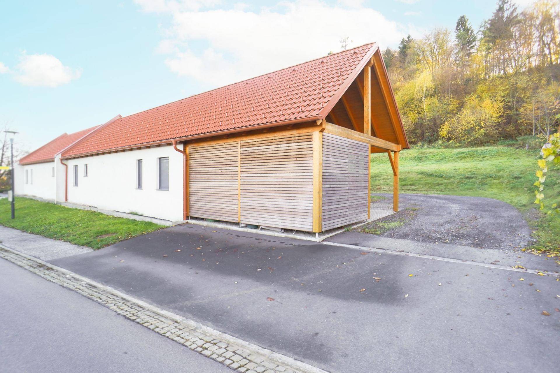 Weißes Gebäude mit Holzverkleidung und überdachtem Stellplatz an einer Zufahrt.