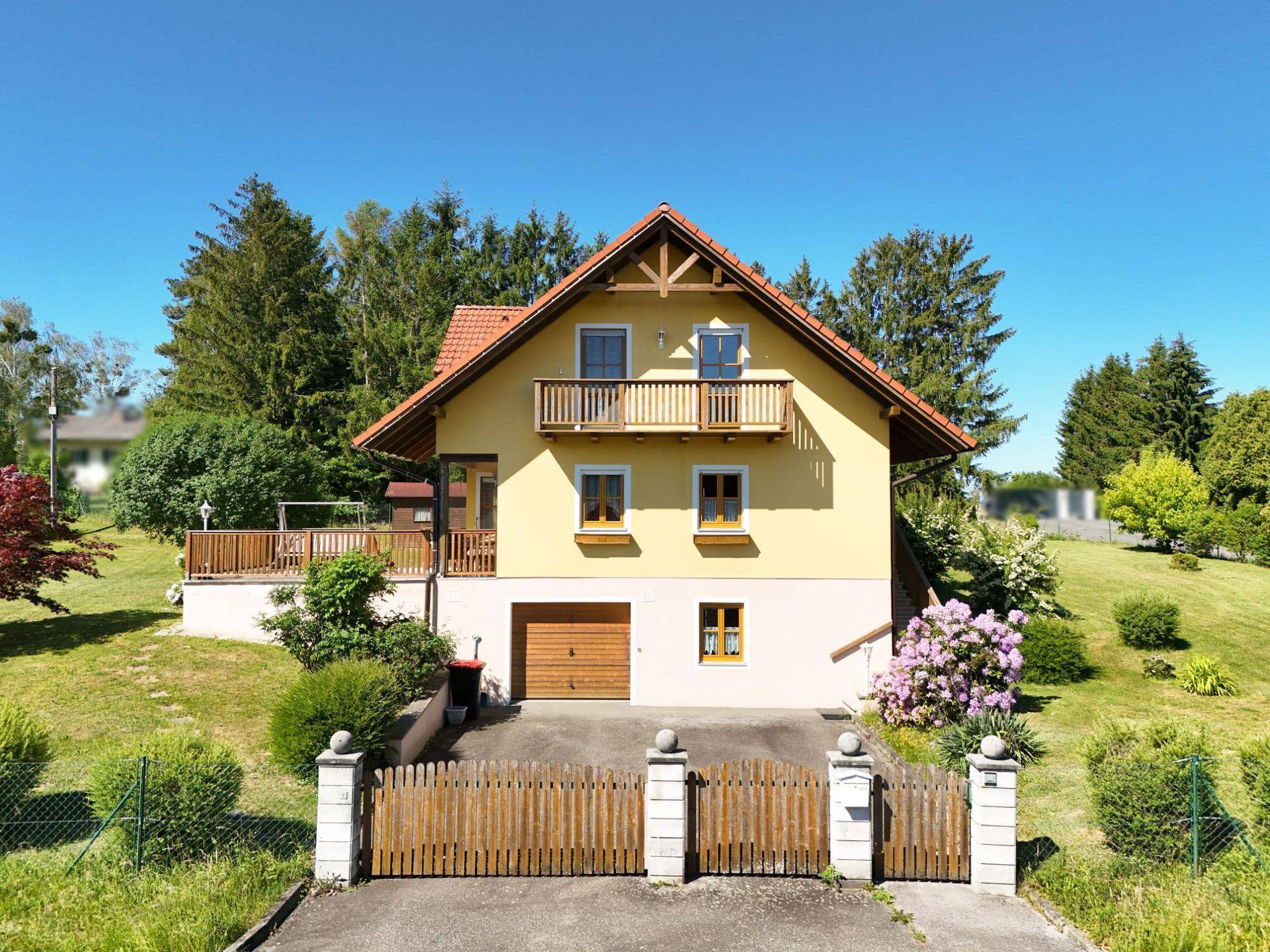 Frontansicht des gelben Hauses mit Holzbalkonen, rotem Dach und einer Einfahrt mit Holztor.