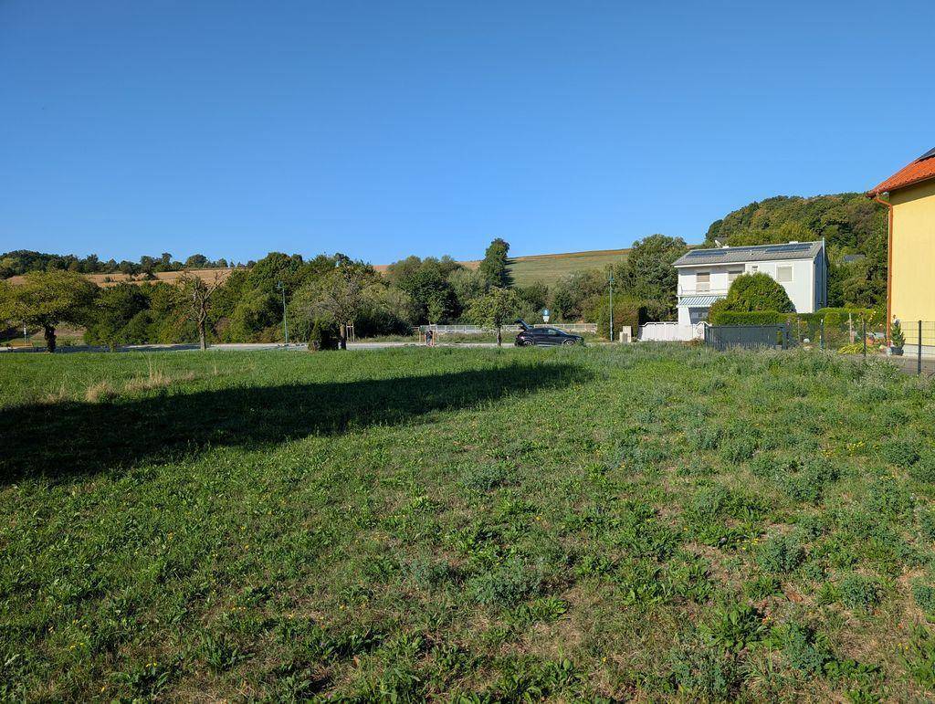 Offenes grünes Grundstück mit Blick auf ein Wohnhaus mit Solaranlage und die umliegende Hügellandschaft.