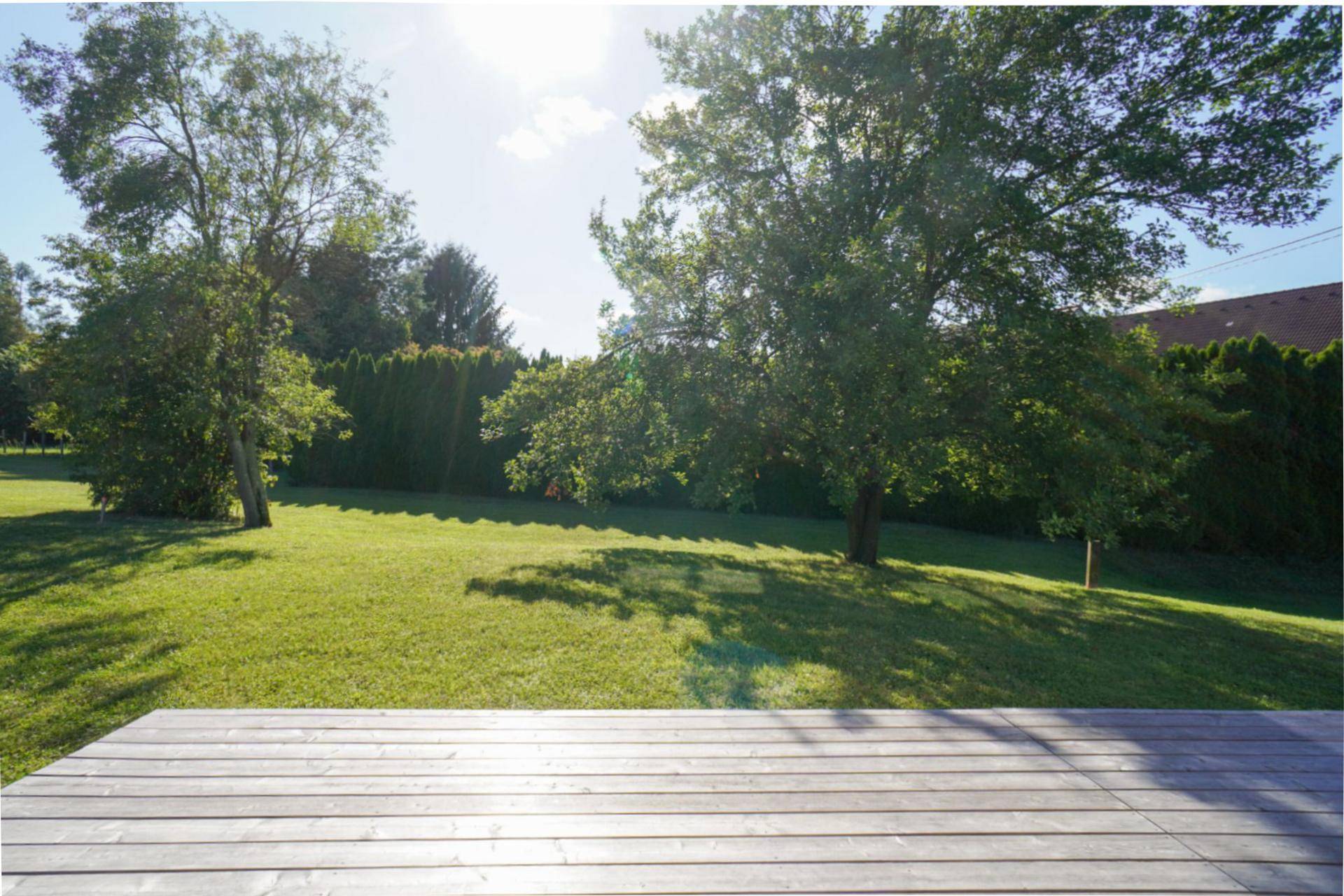 Sonnige Holzterrasse mit Blick auf den weitläufigen grünen Garten und alte Bäume.