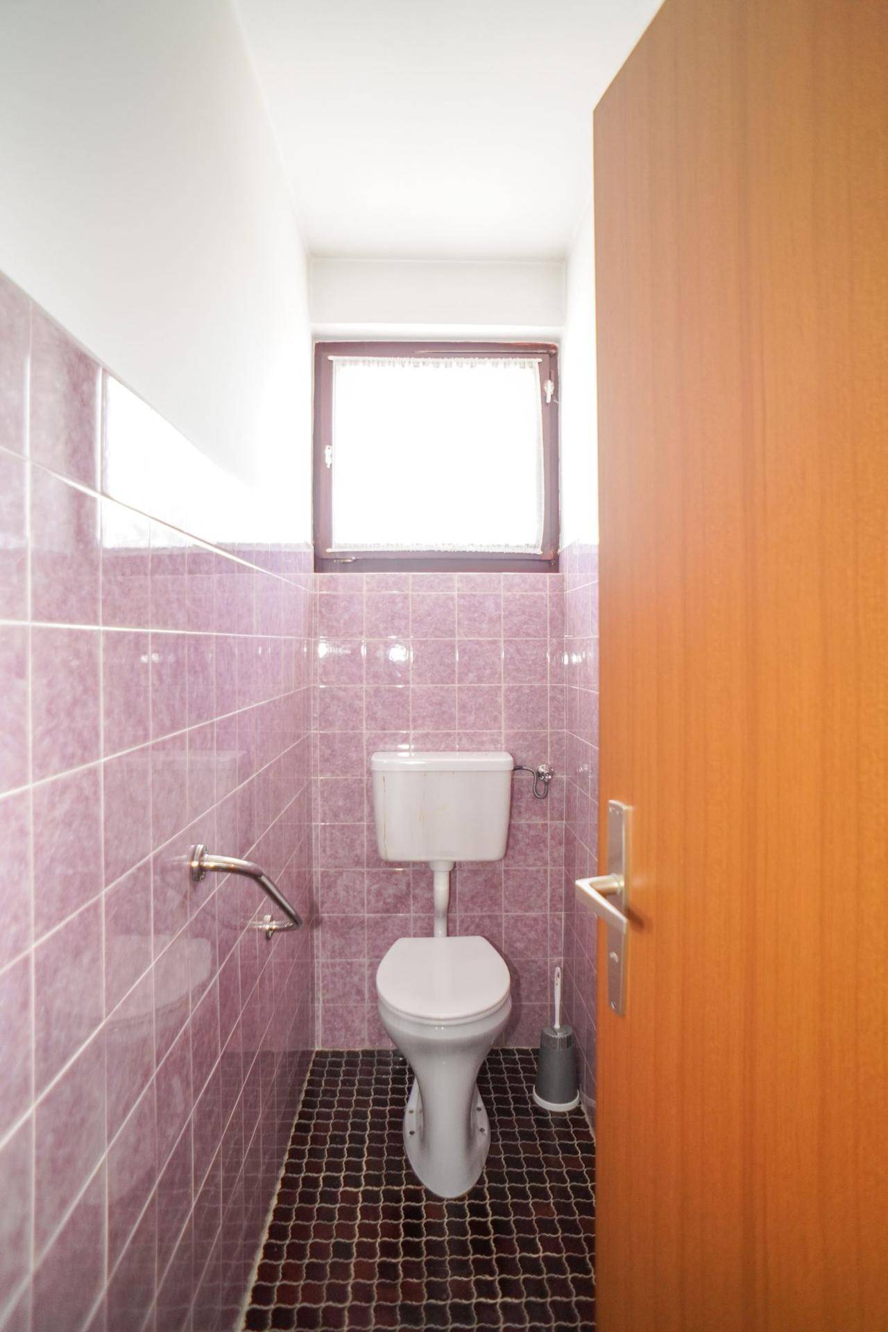 Separate Toilette mit lila Fliesen, Fenster und Haltegriffen für Barrierefreiheit.