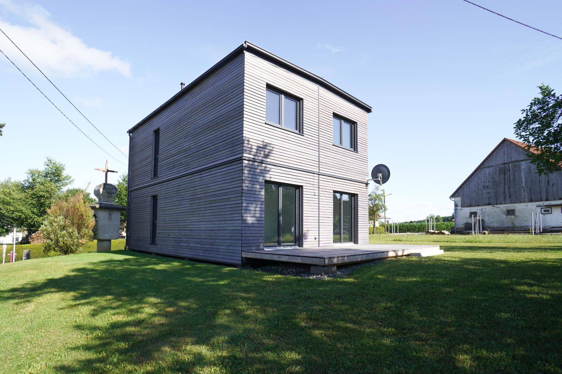 Modernes Einfamilienhaus mit Holzfassade und kleiner Holzterrasse im Grünen unter blauem Himmel.