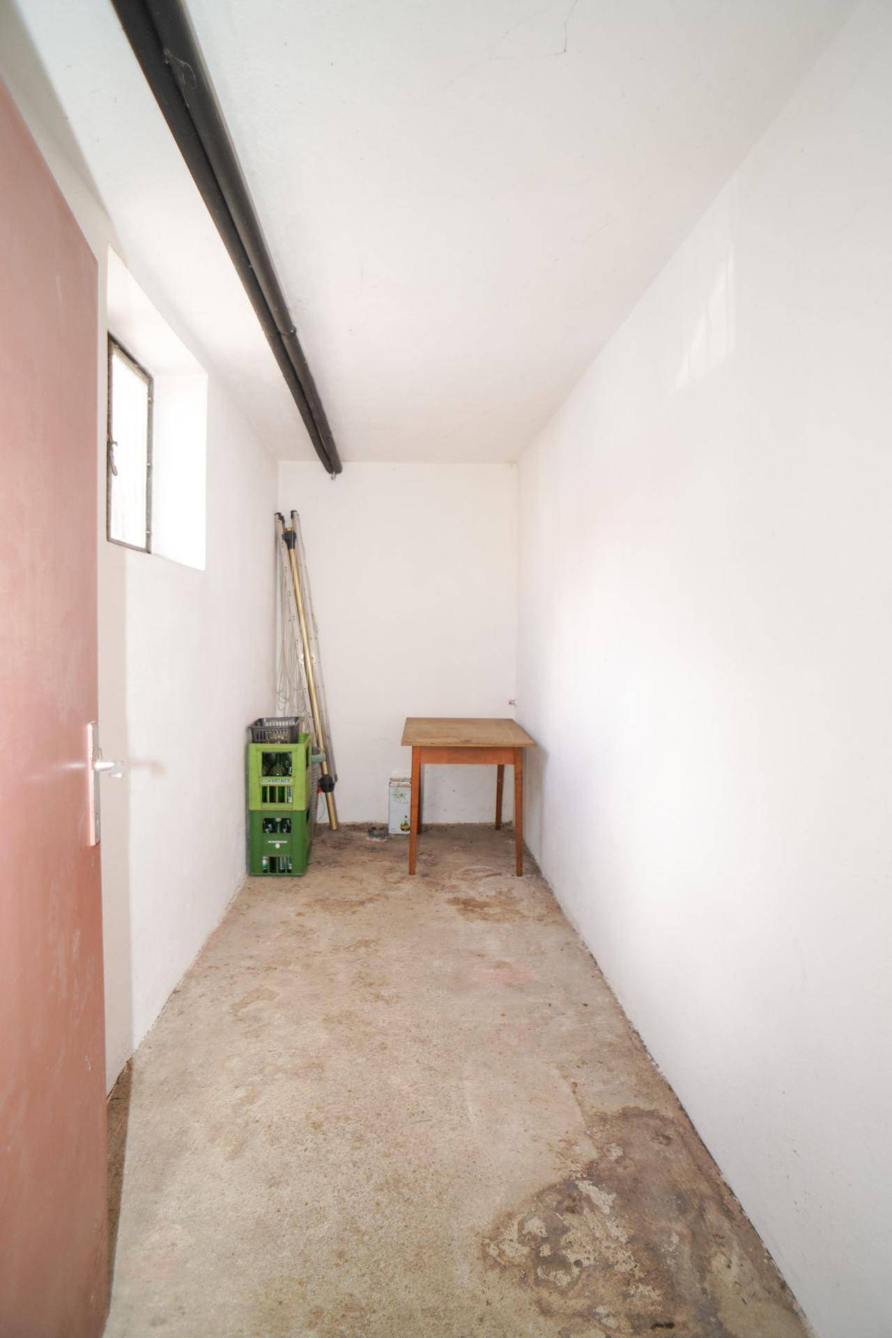 Schmaler Kellerraum mit Betonboden, weiß gestrichenen Wänden und einem kleinen Fenster.